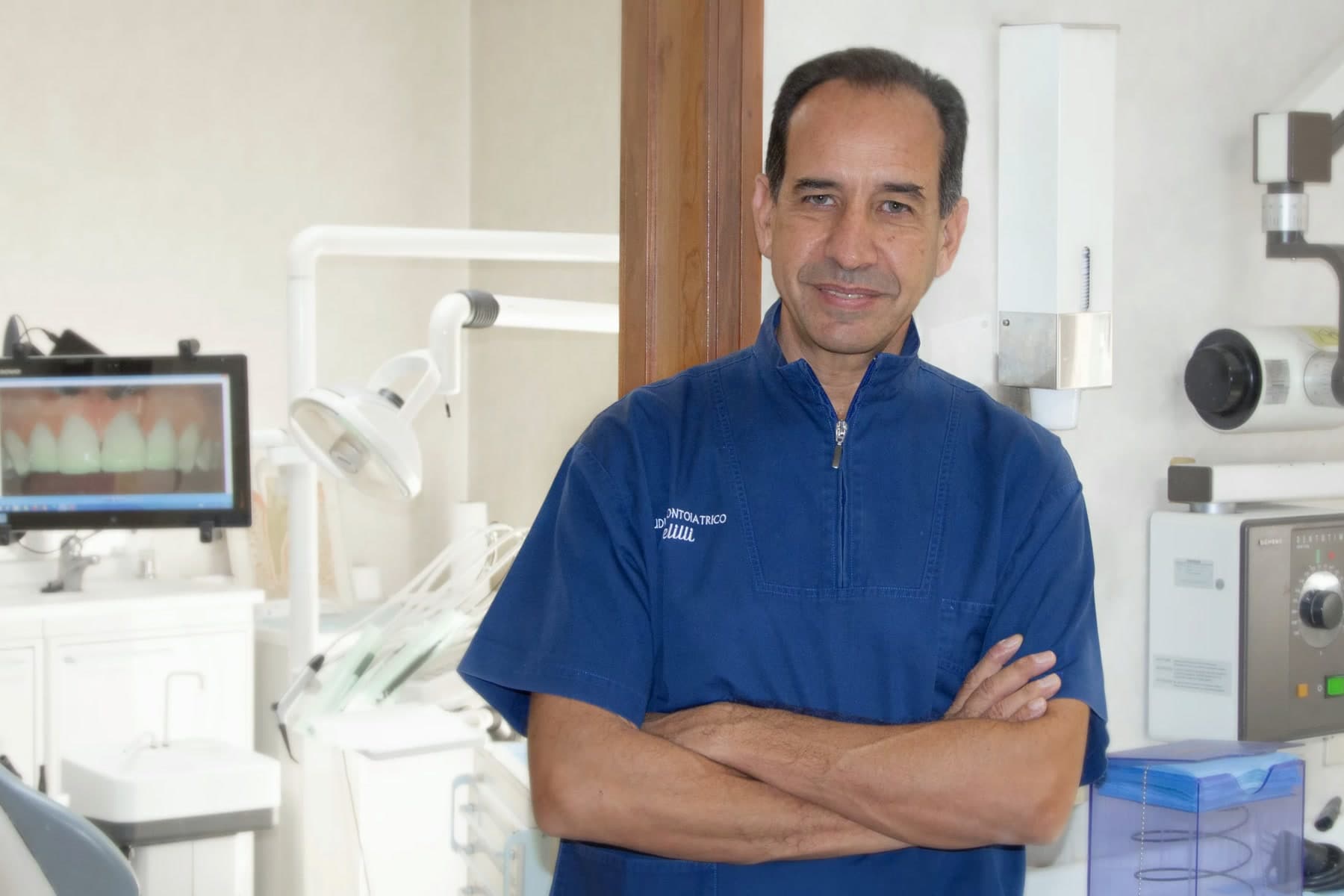 Il dott. Dario Melilli, in divisa blu con il logo dello studio, a braccia conserte nella sala operativa: alle sue spalle il riunito, il monitor con un'immagine dei denti e il radiografico a parete