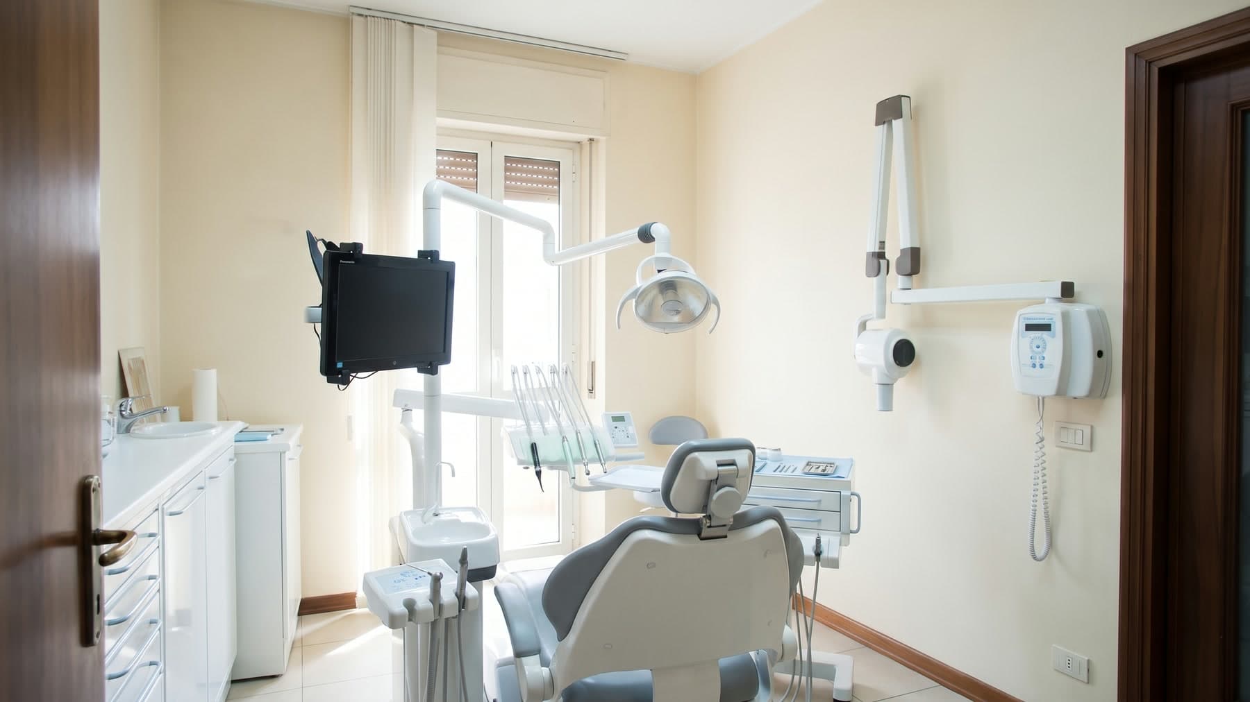 Sala operativa con pareti color crema, stipiti in legno scuro, riunito bianco e grigio, lampada, monitor a braccio e radiografico a parete, finestra con avvolgibili